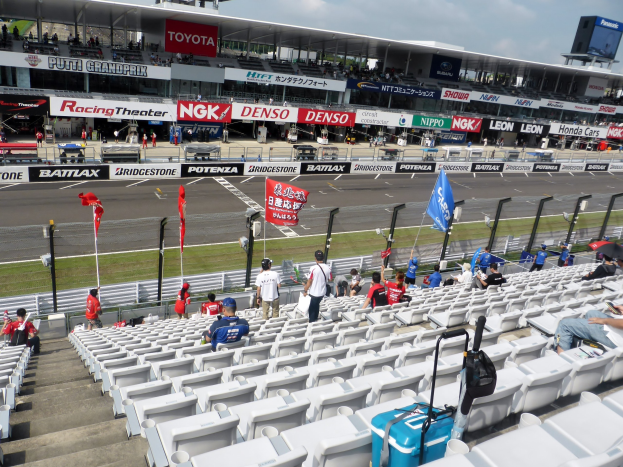Eine Gruppe von Menschen sitzt auf Tribünen in einem Stadion bei einer Rennstrecke, mit Fahnen, Texttafeln, Geländern und Stufen, unter einem bewölkten Himmel.