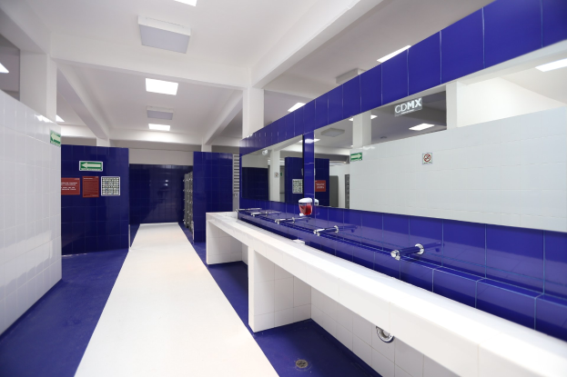 Öffentliche Toilette mit blauen Kachelwänden, weißen Countertops, Waschbecken mit Hähnen, einer Plattform mit einer Flasche und anderen Gegenständen, Informationsschildern an den Wänden, Deckenleuchten und tragenden Säulen.