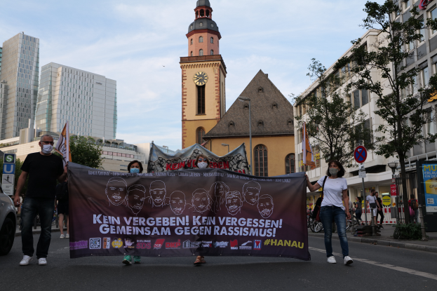 Eine Gruppe von Menschen mit Masken geht eine Straße entlang und hölt ein Plakat, mit einem geparkten Auto auf der linken Seite, Gebäude, Bäume, Schilder, Masten und einen Uhrturm im Hintergrund bei klarem blauem Himmel.