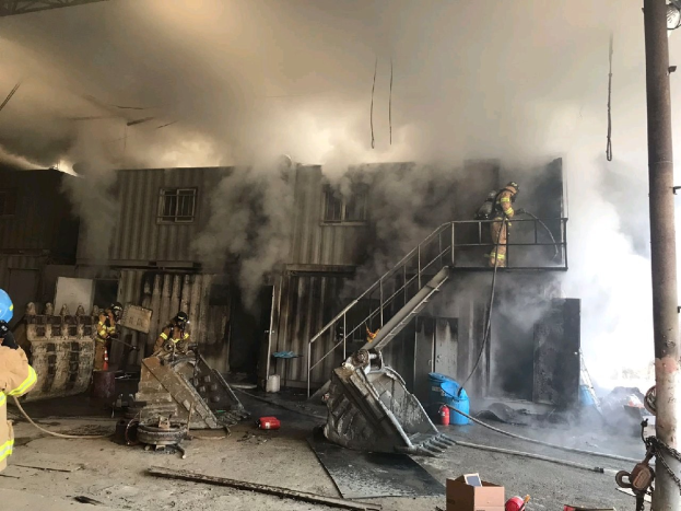 Feuerwehrleute in Helmen und Schutzausrüstung verwenden Schläuche, um ein Lagerfeuer zu löschen, mit Rauch, der aus dem Gebäude aufsteigt, Treppen mit Geländern, die zum Eingang führen, einem Kettenglied-Pfosten, verstreuten Trümmern und Fenstern im Hintergrund.
