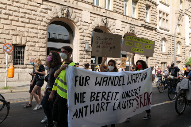 Eine Gruppe von Menschen marschiert auf einer Straße in Berlin, hält Schilder und Banner hoch und fährt mit Fahrrädern, vor einem Gebäude mit Fenstern, Bögen, Säulen, Skulpturen, Bäumen und einem Laternenmast, an einer Anti-Kriegs-Protestaktion teilnehmend.