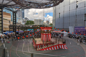 Große Menschenmenge versammelt sich um eine Bühne in einer urbanen Umgebung mit Gebäuden, Bäumen und einem bewölkten Himmel, mit Darstellern und Equipment für die Eröffnung des Tokyo Circus Festivals.