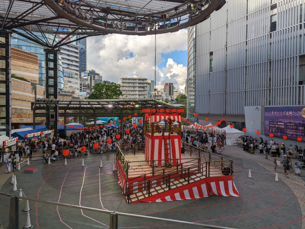 Große Menschenmenge versammelt sich um eine Bühne in einer urbanen Umgebung mit Gebäuden, Bäumen und einem bewölkten Himmel, mit Darstellern und Equipment für die Eröffnung des Tokyo Circus Festivals.