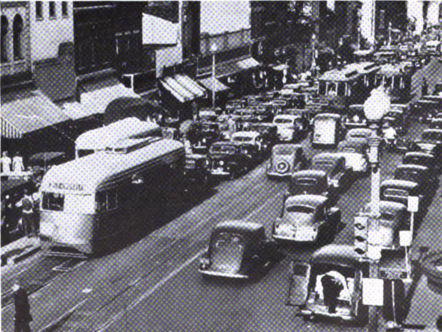 Busy city street with multiple cars, buses, buildings, poles, and pedestrians; text visible at the bottom.