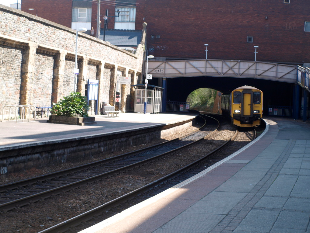 Ein Zug fährt auf den Schienen mit Plattformen auf beiden Seiten, eine Hauswand links, Straßenlaternen, Pflanzen, Bänke, eine Kabine, eine Brücke über die Plattformen und Gebäude und Masten im Hintergrund.