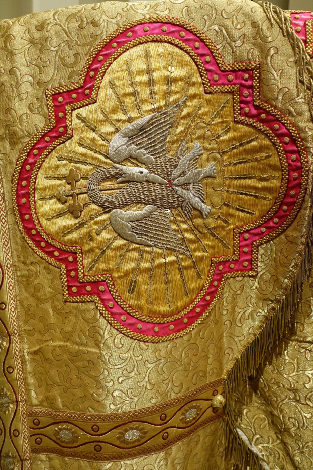 Nahaufnahme eines Priestergewandes mit goldenen und roten Stickereien und detaillierten Mustern, vor einem Wandhintergrund.