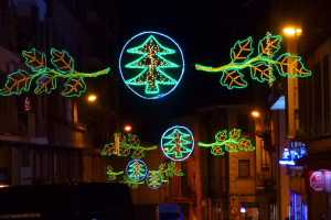 Eine städtische Straße, geschmückt mit festlichen Weihnachtslichtern mit Blättern und anderen Schmuckelementen, mit Fahrzeugen, die an Gebäuden auf beiden Seiten vorbeifahren.