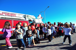 Gruppe von Kindern vor einem Feuerlöschfahrzeug mit der Aufschrift Yokosuka Feuerwehr während eines Weihnachtsumzugs auf einer Straße mit Laternenmasten, Bäumen und einem klaren blauen Himmel.