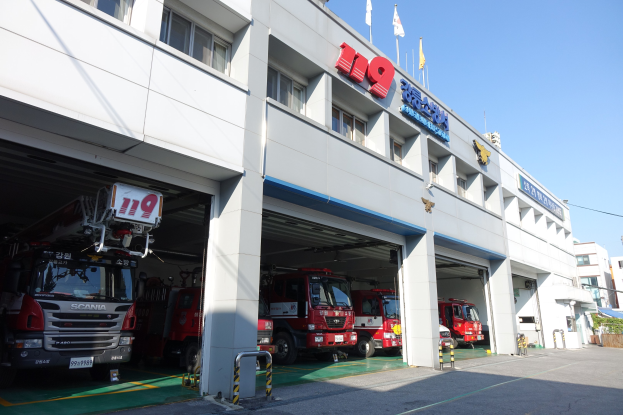 Feuerwache mit mehreren Feuerwehrautos davor, mit Fenstern, Säulen, Namensschildern und Flaggen mit Stangen im Vordergrund, vor anderen Gebäuden unter einem klaren blauen Himmel.