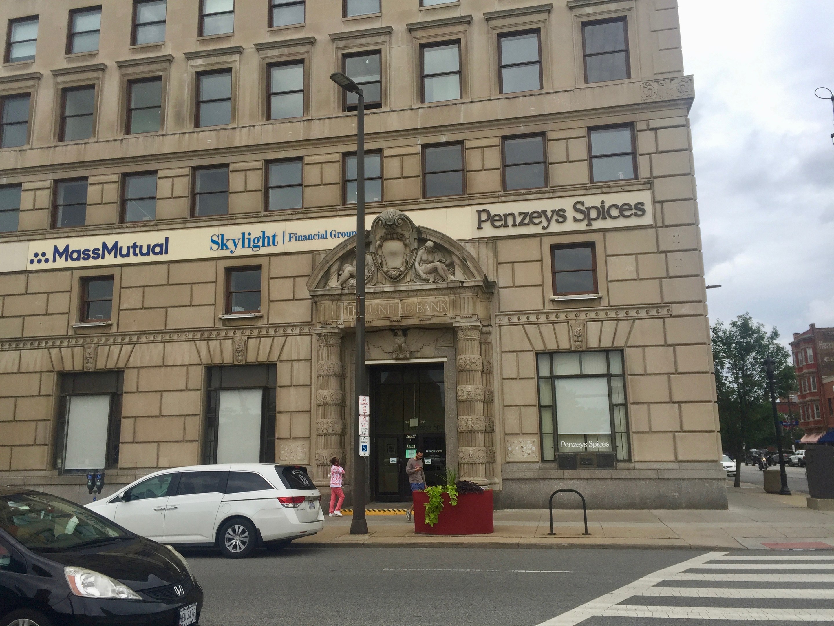 Großes Gebäude mit parkenden Autos davor, Straßenlaternen, bewegliche Fahrzeuge, Fußgänger auf dem Gehweg, Grünfläche und bewölkter Himmel, identifiziert als Hauptsitz der Massachusetts Mutual Financial Group.