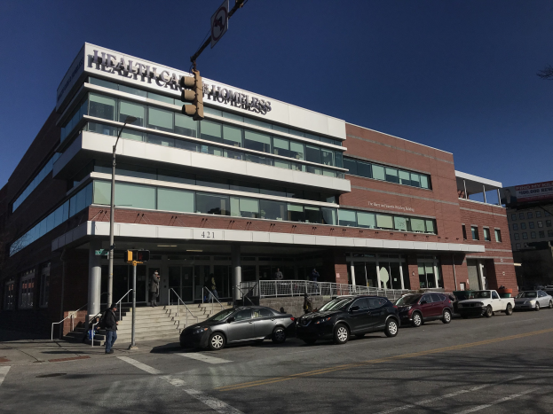 Großes Gebäude mit der Aufschrift "New York City Health Care Homeless Center" mit parkenden Autos davor, umgeben von Straßeninfrastruktur, Fußgängern, Bäumen und einem klaren blauen Himmel.