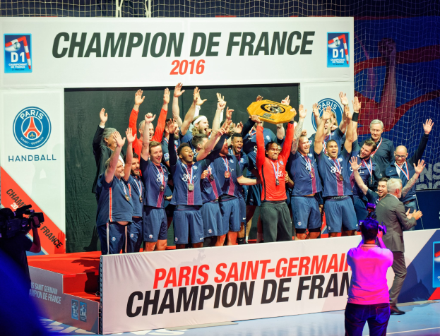 Eine Gruppe von Männern hält einen Pokal auf einer Bühne, während eine Person filmt und ein 'Champion de France 2016'-Schild im Hintergrund zu sehen ist.