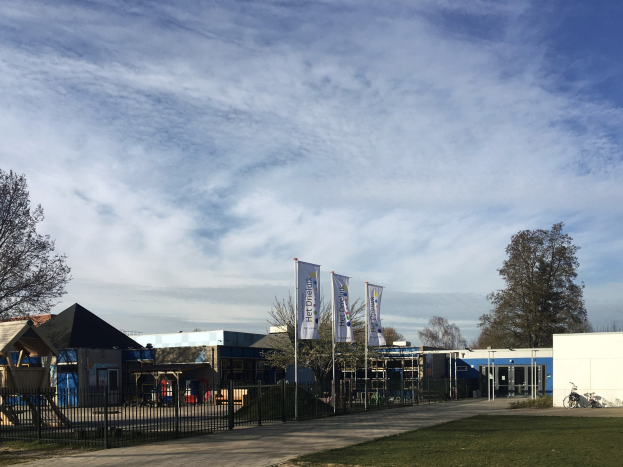 Großes Gebäude mit einem Metallzaun, Rasenfläche, Weg, Bäumen, Bannern, einem Fahrrad, nahen Häusern und einem bewölkten Himmel, identifiziert als der neue Spielplatz der Schule.