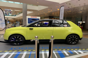 Ein gelbes wasserstoffbetriebenes Elektroauto steht vor einem Gebäude, umgeben von Absperrpollern mit Seilen, Bannern und Schildern, mit einer Person im Auto, einer Gruppe von Menschen in der Nähe, einer Säule, einem Topfpflanze, Deckenleuchten und einer Decke mit Deckenventilatoren.