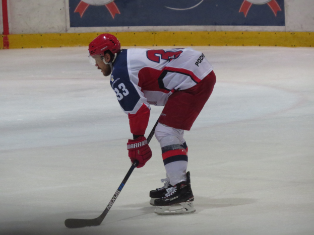 Eishockey-Spieler in voller Ausrüstung auf dem Eis mit einem Stock, der einer Wand mit Banner im Hintergrund gegenübersteht.