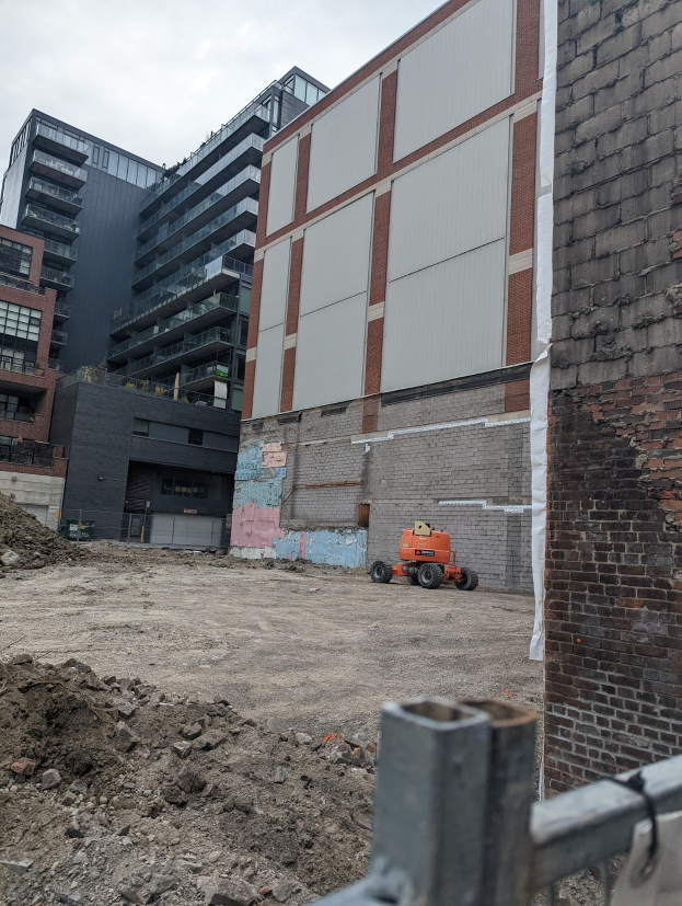 Baustelle mit einem großen Gebäude im Hintergrund, das abgerissen wird, ein Fahrzeug auf dem Boden, ein Metallzaun und ein bewölkter Himmel.