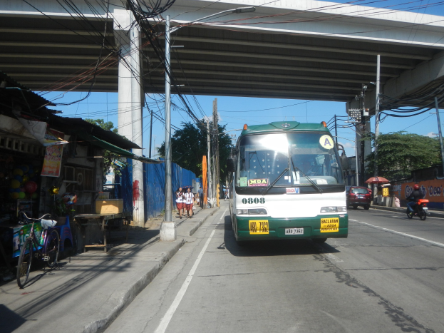 Ein grüner und weißer Bus fährt auf einer Straße neben einer Brücke, mit Strommasten und -leitungen entlang der Straße, Fußgängern auf dem Gehweg, parkenden Fahrrädern und Geschäften an der Straße, unter einem klaren blauen Himmel mit Bäumen im Hintergrund.