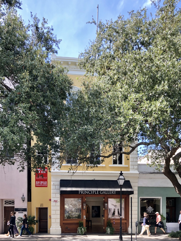 Außenansicht der Principle Gallery in Charleston, SC, mit Menschen auf dem Bürgersteig, Topfpflanzen, einer Straßenlaterne, einem Schild, einer Fahne mit Text und einem Wolkenhimmel.
