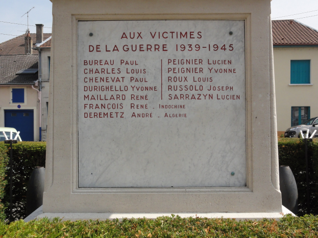 Denkmal mit der Inschrift "Aux Victimes de la Guerre 1939-1945" vor einem Gebäude, umgeben von Pflanzen, Fahrzeugen, Versorgungsmasten und Häusern unter einem bewölkten Himmel.