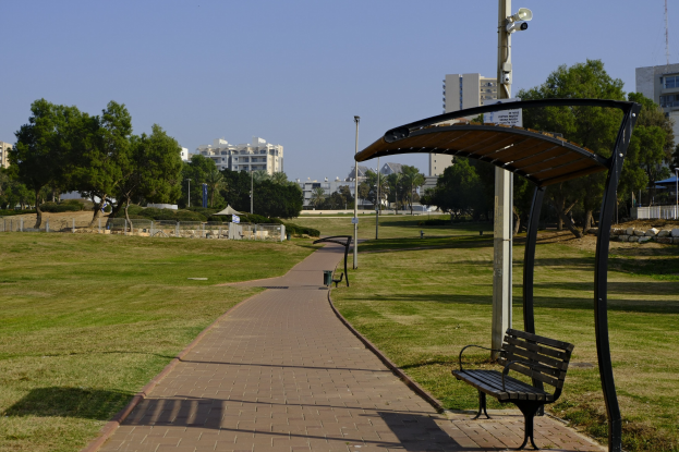 Eine Parkbank auf einem grünen Feld mit Bäumen, Masten, Lichtern und Gebäuden im Hintergrund und einem Mülleimer im Vordergrund.